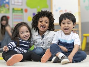 Three children sitting on the floor of a daycare classroom smiling and laughing.