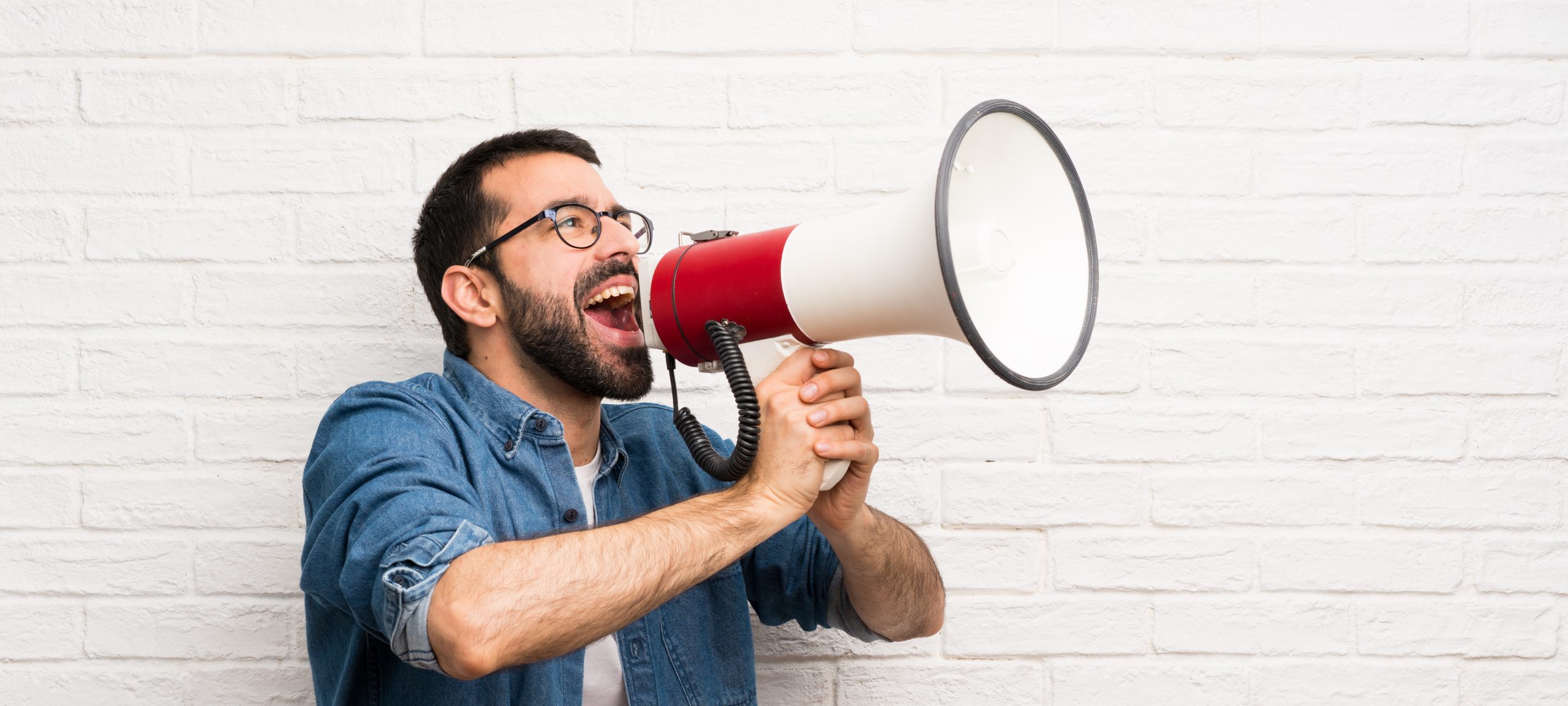 Handsome man with beard over white brick wall shouting through a megaphone