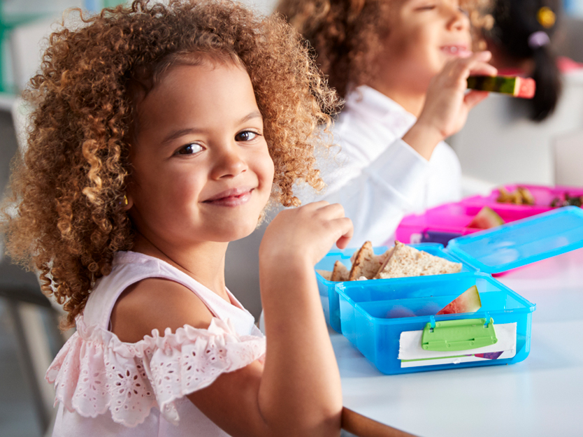 Image of young girl smiling while eating lunch from lunch box at a table with other children in the background.