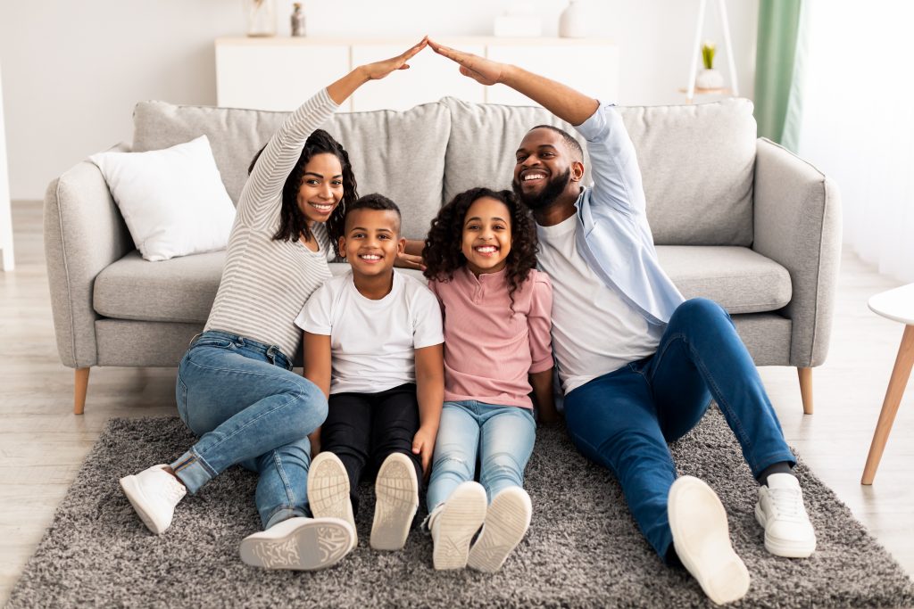 Family Care, Protection And Insurance Concept. Portrait of smiling African American parents making symbolic roof of hands above their happy children, sitting on the floor carpet in living room at home