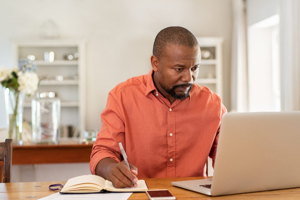 Mature man working on laptop while taking notes. Businessman working at home with computer while writing on agenda. African man managing home finance, reviewing bank account and using laptop in living room.