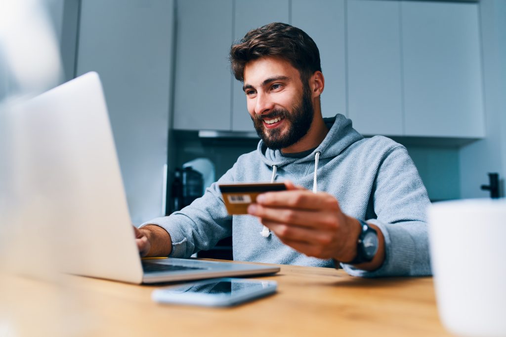 Cheerful young man paying bills online with credit card and laptop